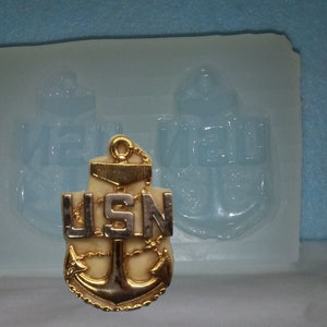 May include: A gold and silver USN anchor pin. The pin features a gold anchor with a chain and the letters "USN" in silver. The pin is set against a clear mold with two anchor impressions. The background is a light blue.