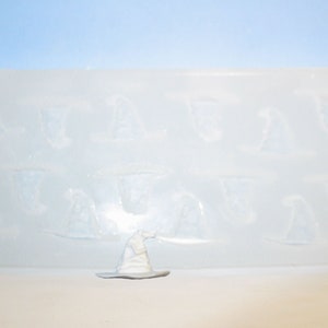 May include: A clear silicone mold with 12 impressions of a wizard's hat. The mold is on a white surface with a white wizard's hat in the foreground.