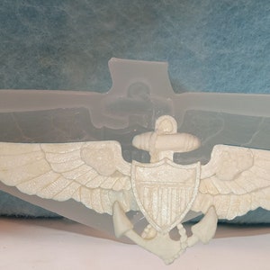 May include: A white silicone mold of a Naval Aviator wings and anchor insignia. The mold is designed to create a 3D replica of the insignia.