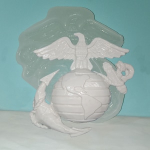 May include: A white plastic mold of the United States Marine Corps emblem. The emblem features an eagle perched atop a globe, with an anchor wrapped around the globe.