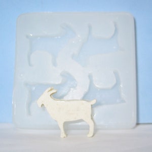 May include: A white goat figurine next to a clear silicone mold with goat-shaped cavities. The mold is square and has a smooth surface. The goat figurine is standing upright.