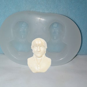 May include: A light-colored bust of a person with short hair and a collared shirt sits in front of a clear silicone mold. The mold has two bust impressions. The background is a light blue.
