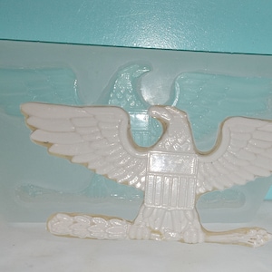 May include: A white plastic mold of an eagle with outstretched wings, a shield on its chest, and a branch in its talons. The mold is on a clear silicone mat.
