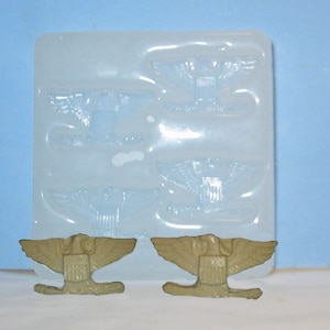 May include: Two gold-colored eagle insignia with wings spread, each with a shield on the chest. The insignia are sitting on a white silicone mold with four impressions of the same insignia.