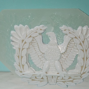 May include: A white plastic mold of an eagle with wings spread, surrounded by a wreath of leaves. The eagle is a symbol of the United States Army.