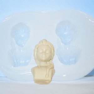 May include: A white silicone mold with two impressions of a girl's head and one impression of a girl's bust. The mold is on a white surface.