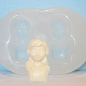May include: A white silicone mold with two impressions of a person's head and shoulders, and one impression of a person's bust. The mold is on a light blue surface.