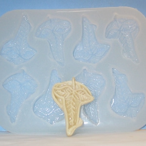 May include: A clear silicone mold with eight impressions of a leaf design. The mold is on a blue surface and a white leaf-shaped object is in front of the mold.