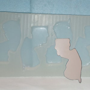 May include: A clear silicone mold with multiple cavities shaped like the state of New Jersey. One cavity is filled with a light pink substance.