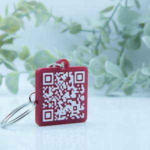 May include: A red square keychain with a white QR code design. The keychain has a silver key ring attached. The background features light green foliage against a white surface.