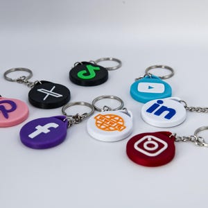 May include: A set of nine keychains featuring various social media logos. The keychains are made of plastic and have a round shape. The logos include Pinterest, Facebook, a globe with www. on it, Instagram, LinkedIn, YouTube, TikTok, and a black and white cross.