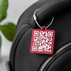 May include: A square red luggage tag featuring a white QR code pattern on its front surface. The tag is attached to a black bag handle by a thin, flexible silver-colored metal cable loop with a small screw-type fastener at the top.