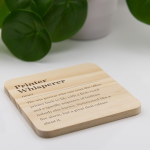 May include: A square light-colored wooden coaster featuring the printed text Printer Whisperer, noun, and a definition describing a person who fixes office printers with specific button sequences, presented in a dark brown sans-serif font.