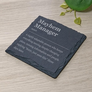 Könnte beinhalten: Ein quadratischer Untersetzer aus dunkelgrauem Schiefer mit rauen, unebenen Kanten. Die Oberfläche ist mit weißem Text graviert: Mayhem Manager, noun, A highly adaptable person who keeps everything moving despite changing plans, missing information and people replying "Sorry, just seen this" three days later.