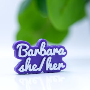 May include: A purple name tag with the name "Barbara" and the pronouns "she/her" in white cursive lettering. The tag is set against a white surface, with a blurred green plant visible.