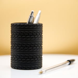 May include: A black pen holder with a tyre tread design holds two silver pens. The holder is on a white surface.