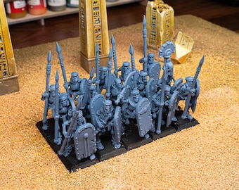 May include: A group of gray miniature skeleton warriors, armed with spears and shields, stand on a black base. The figures are detailed and appear to be part of a tabletop game or model set. The background includes miniature obelisks.