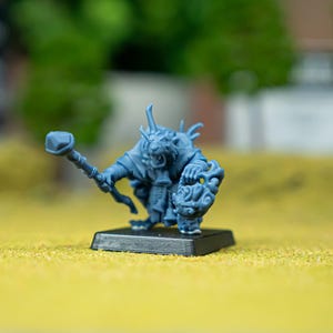 Op de afbeelding: Een grijs, onbeschilderd miniatuurfiguur van een ratachtig wezen op een zwarte vierkante voet. Het figuur houdt een staf met een steenvormige top in de ene hand en een rond, versierd schild in de andere, met gedetailleerde texturen van vacht en kleding.
