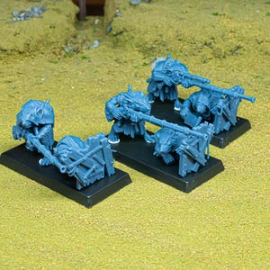 Peut inclure: Trois figurines miniatures en plastique bleu non peintes représentant des créatures rats anthropomorphes, chacune montée sur une base rectangulaire noire. Les personnages tiennent de longs instruments semblables à des armes, derrière des barricades.