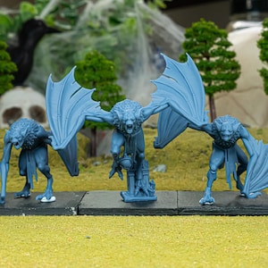 Puede incluir: Tres figuras de monstruos azul-grisáceos con alas de murciélago y rostros amenazantes se alzan sobre una plataforma oscura. Las figuras tienen cuerpos humanoides y están posadas en una postura amenazante. El fondo incluye árboles en miniatura, una calavera y un cuervo negro.