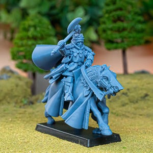 Puede incluir: Figura en miniatura azul de un caballero a caballo, sosteniendo un arma. El caballero está blindado y el caballo está adornado con una armadura detallada. La figura está montada sobre una base negra, sobre un fondo de follaje verde y una superficie herbosa.