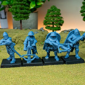 May include: Four blue miniature figurines of medieval soldiers with crossbows, standing on black bases. The figures are detailed with helmets, armor, and weapons, set against a backdrop of miniature trees and a grassy surface.