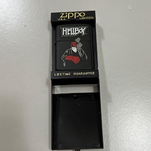 May include: Black Zippo lighter with a Hellboy graphic. The lighter is in a black display case. The lighter features the text "Hellboy" and an image of the character. The display case has the text "Zippo U.S.A. Lighter" and "Lifetime Guarantee".