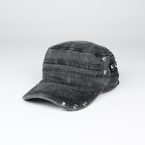 Könnte beinhalten: Eine militärisch anmutende Kappe aus verwaschenem, anthrazitfarbenem Denim. Die Mütze hat eine flache Oberseite, einen gebogenen Schirm mit silberfarbenen Metallösen am Rand und ein seitliches Riemendetail, das mit passenden Metallnieten befestigt ist.