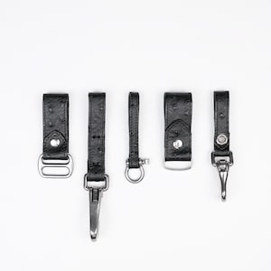 Könnte beinhalten: Fünf schwarze Schlüsselanhänger aus strukturiertem Leder, die in einer Reihe angeordnet sind. Jeder Riemen verfügt über silberfarbene Metallbeschläge, darunter verschiedene Clips, D-Ringe und Druckknöpfe. Das Leder hat ein punktiertes Muster.