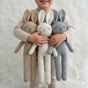 Puede incluir: Tres muñecos de conejito de ganchillo en beige, blanco y gris. Cada conejito tiene orejas largas, ojos cerrados y una cara sencilla. Los muñecos son suaves y achuchables, perfectos para niños.