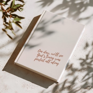 Peut inclure: Un journal crème avec le texte "One day, we'll see God's timing was perfect all along" en script marron. Le journal est posé sur une surface blanche avec des ombres de feuilles.