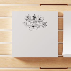 May include: A white notepad with a floral design and the text "WHAT IS ON GOD'S HEART FOR YOU TODAY?" in a rectangular frame. The notepad is on a wooden surface.
