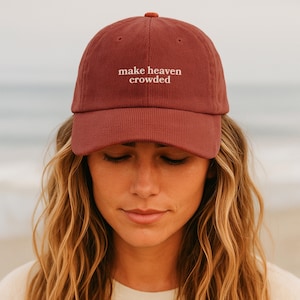 Puede incluir: Gorra de béisbol de pana granate con la frase "make heaven crowded" bordada en blanco. La gorra tiene una visera curva y un botón a juego en la parte superior. La persona lleva el pelo largo y rubio ondulado.