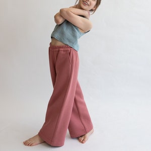 Puede incluir: Una persona joven que lleva una blusa sin mangas de color azul claro y pantalones de color rosa de piernas anchas. Los pantalones tienen una cintura con cordón y bolsillos.