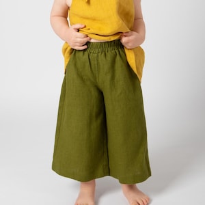 May include: A young child wearing a yellow linen top with a tied neckline and olive green linen trousers.