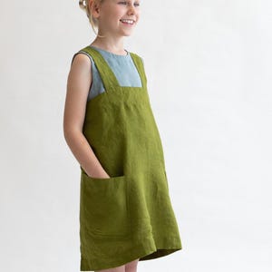 Puede incluir: Una persona joven lleva un vestido de delantal de lino verde con un top sin mangas azul claro debajo. El vestido tiene un bolsillo grande en la parte delantera y un lazo blanco en los calcetines.