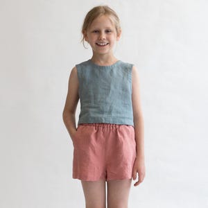 May include: A young person wearing a light blue sleeveless linen top and coral linen shorts. The shorts have an elastic waistband and are pulled up to the waist. The person is wearing white socks and white sneakers.