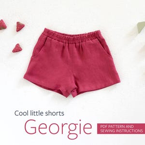 Könnte beinhalten: Rote Shorts mit Taschen, beschriftet mit "Georgie" und "Cool little shorts". Die Shorts liegen auf einer weißen Oberfläche mit erdbeerförmigen Dekorationen und geometrischen Formen.