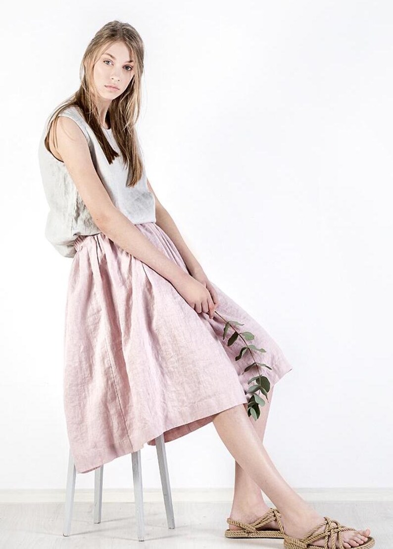 May include: A woman wearing a light gray sleeveless top and a pink linen skirt. She is sitting on a white stool and holding a sprig of greenery.