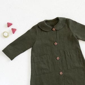 Puede incluir: Una chaqueta de lino verde oscuro con cuello de Peter Pan y dos bolsillos. La chaqueta tiene botones marrones.