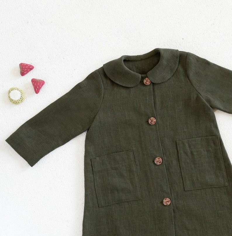 Puede incluir: Una chaqueta de lino verde oscuro con cuello de Peter Pan y dos bolsillos. La chaqueta tiene botones marrones.