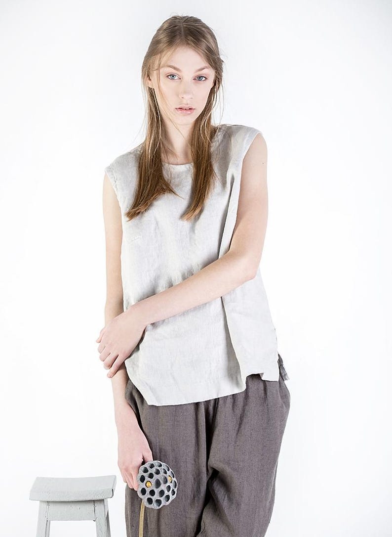 May include: A woman wearing a light gray sleeveless linen top and brown linen pants. She is holding a dried lotus pod in her hand.