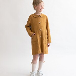 Puede incluir: Un vestido de lino amarillo mostaza con cuello de Peter Pan y cierre de botones. El vestido tiene dos bolsillos de parche y lo lleva puesto una persona joven con calcetines blancos y zapatillas deportivas.