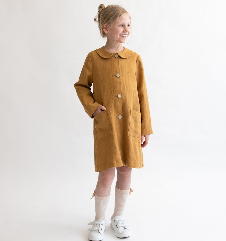 Puede incluir: Un vestido de lino amarillo mostaza con cuello de Peter Pan y cierre de botones. El vestido tiene dos bolsillos de parche y lo lleva puesto una persona joven con calcetines blancos y zapatillas deportivas.