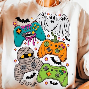 Puede incluir: Una sudadera crema con un gráfico de Halloween. El diseño incluye mandos de juego de dibujos animados en verde azulado, naranja y verde, junto con un fantasma, una momia y murciélagos. Los mandos tienen detalles únicos y espeluznantes.