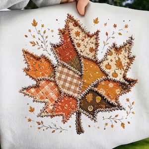 May include: A white sweatshirt featuring a large, central graphic of a maple leaf constructed from various fabric patches. The leaf design incorporates orange, brown, and cream patterns including plaid, pumpkins, and foliage, with frayed edges.