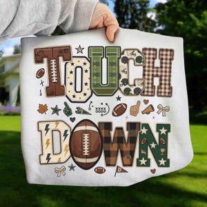 May include: A white t-shirt featuring the word TOUCHDOWN in large, decorative block letters. The letters incorporate various patterns including football textures, plaid, stars, and lightning bolts. Small football-themed icons are scattered throughout.