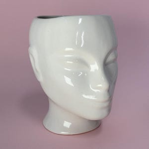 Può includere: Un vaso bianco in ceramica a forma di testa con occhi chiusi, naso e labbra. Il vaso ha una finitura liscia e lucida e poggia su uno sfondo rosa. La testa è un oggetto decorativo.