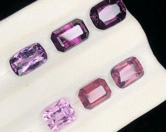 Può includere: Un assortimento di pietre preziose sfaccettate in varie tonalità di viola e rosa. Le gemme sono rettangolari e ovali, con sfaccettature interne visibili. Sono esposte su una superficie bianca, con uno sfondo scuro.