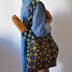 May include: A woman is carrying a blue and yellow patterned tote bag with a baguette inside. The bag has a large pocket on the front with a different pattern.